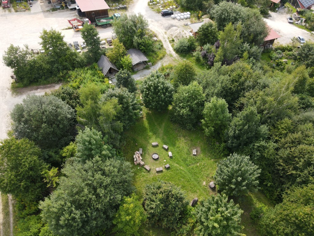 Luftaufnahme eines grünen Geländes mit einem steinernen Sitzkreis, umgeben von Bäumen und Sträuchern, in der Nähe von Gebäuden und Wegen.